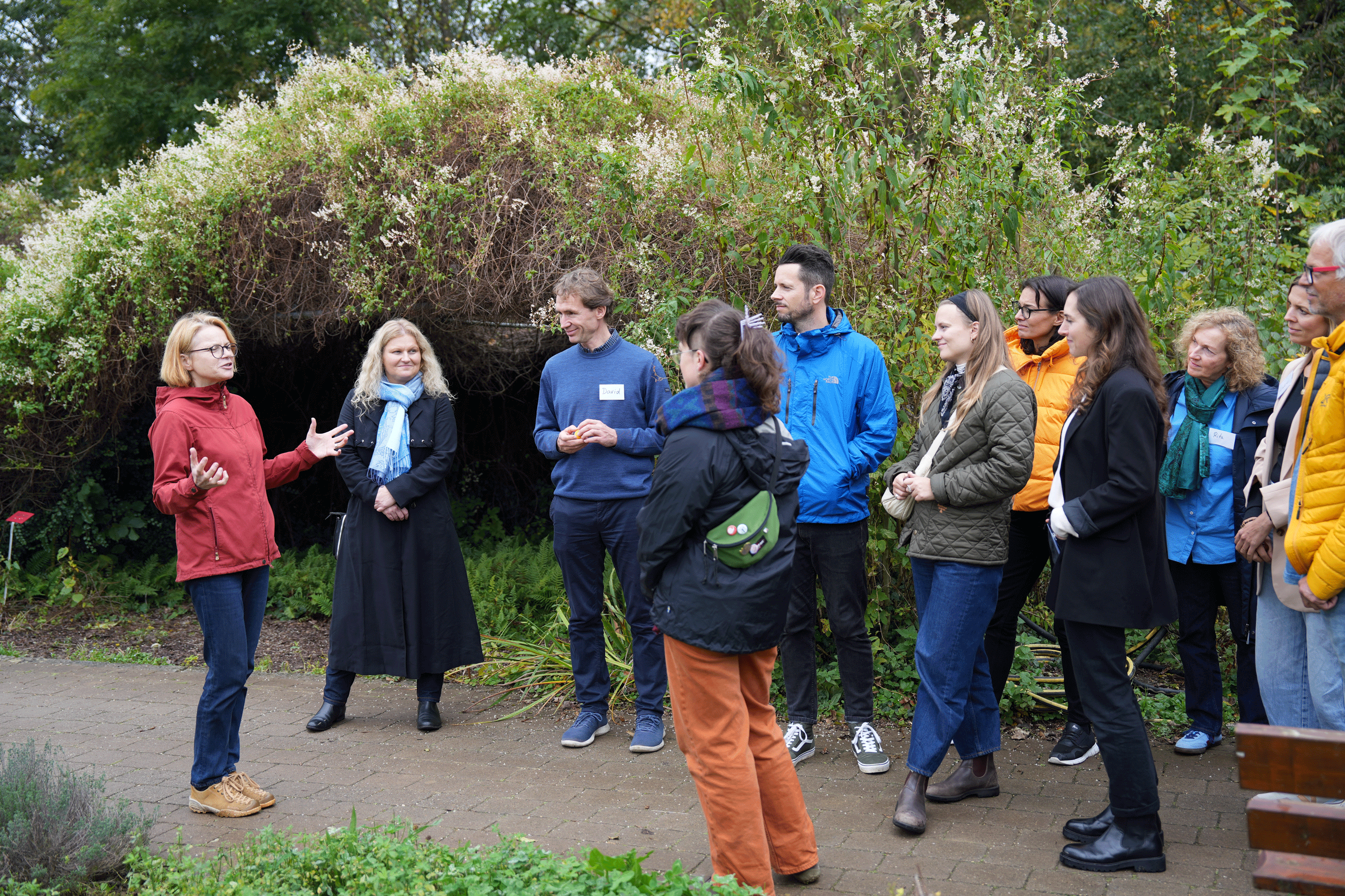 Eine Gruppe von etwa einem Dutzend Menschen steht im Halbkreis im Freien. Eine Frau spricht, w&auml;hrend die anderen aufmerksam zuh&ouml;ren. Im Hintergrund: ein mit Pflanzen bewachsener Laubtunnel.