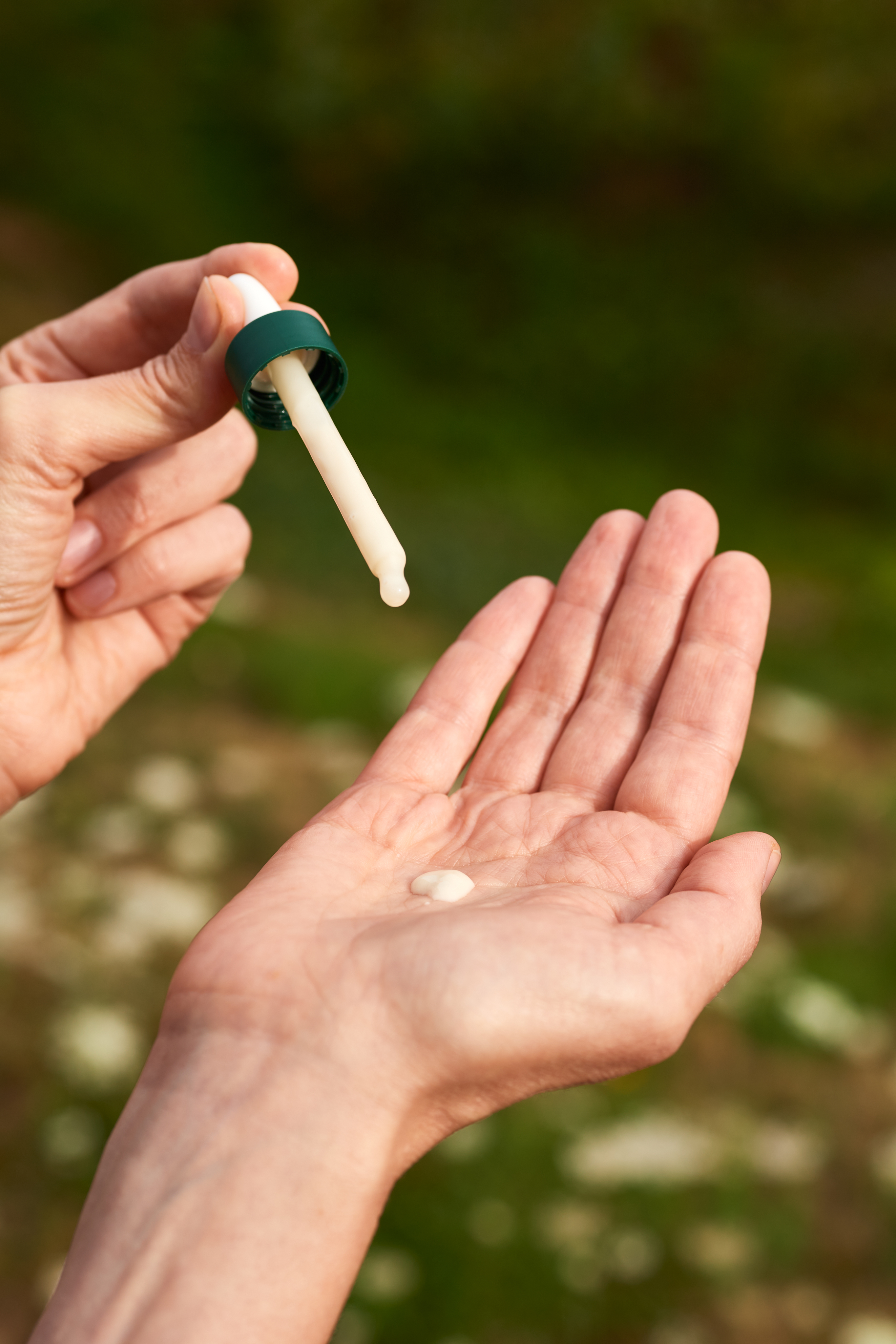 Eine Hand h&auml;lt eine Pipette, aus der eine kleine Menge wei&szlig;er Creme in die ge&ouml;ffnete Innenfl&auml;che der anderen Hand getropft wird. Der Hintergrund ist unscharf und gr&uuml;n.