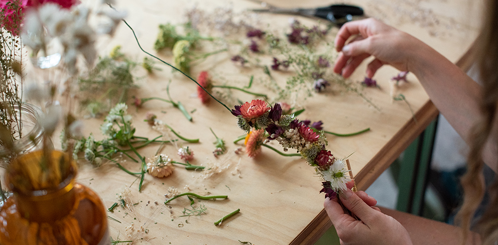 Eine Person fertigt einen Kranz aus Trockenblumen an. Auf dem Tisch liegen verschiedene Bl&uuml;ten, St&auml;ngel und Werkzeuge. Die H&auml;nde formen den Kranz aus den gesammelten Materialien.