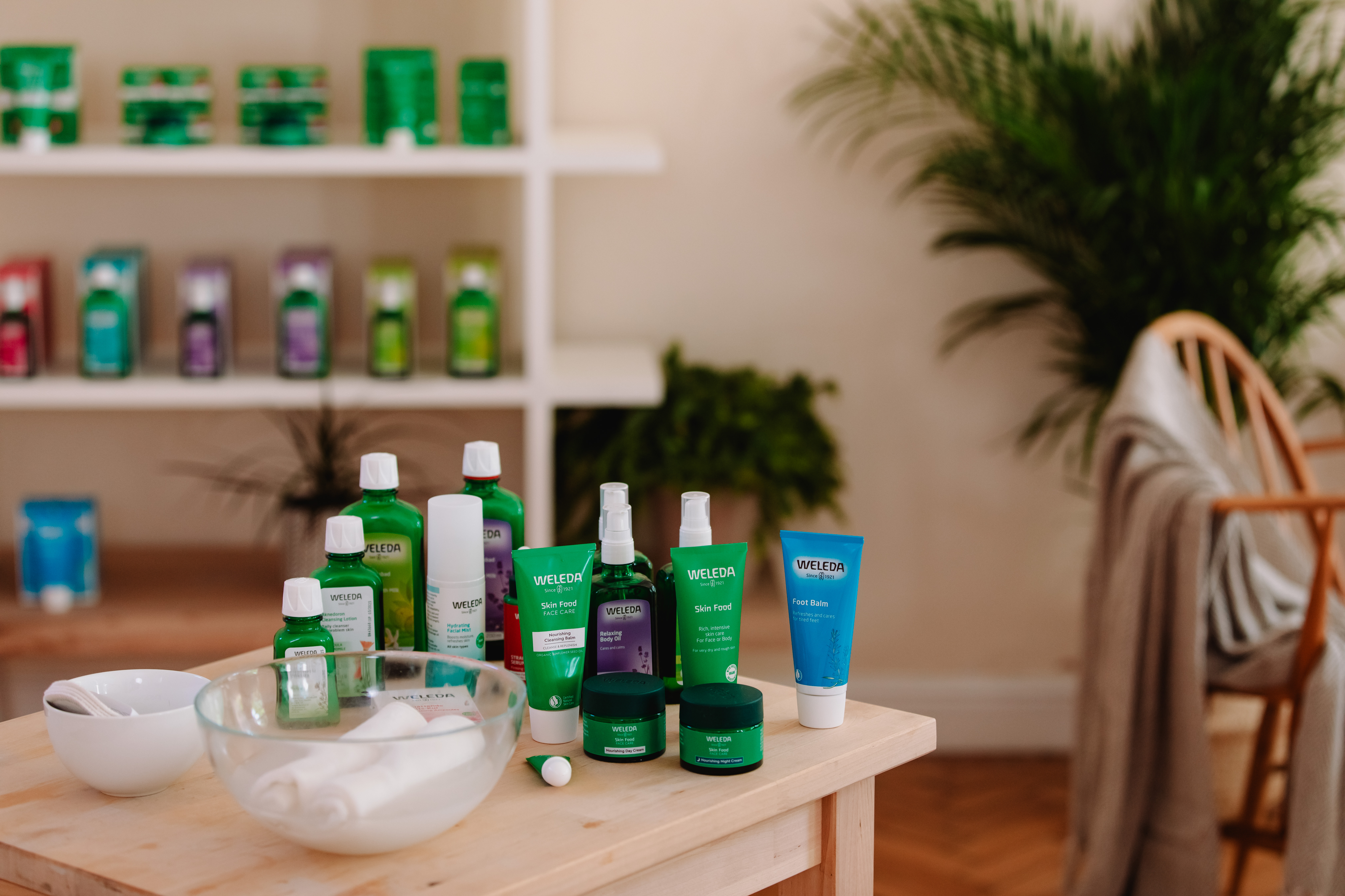 Pflegeprodukte von Weleda auf einem Holztisch, darunter Skin Food, &Ouml;le und Balsame. Im Hintergrund ein Regal mit weiteren Produkten sowie Zimmerpflanzen und ein Holzstuhl.