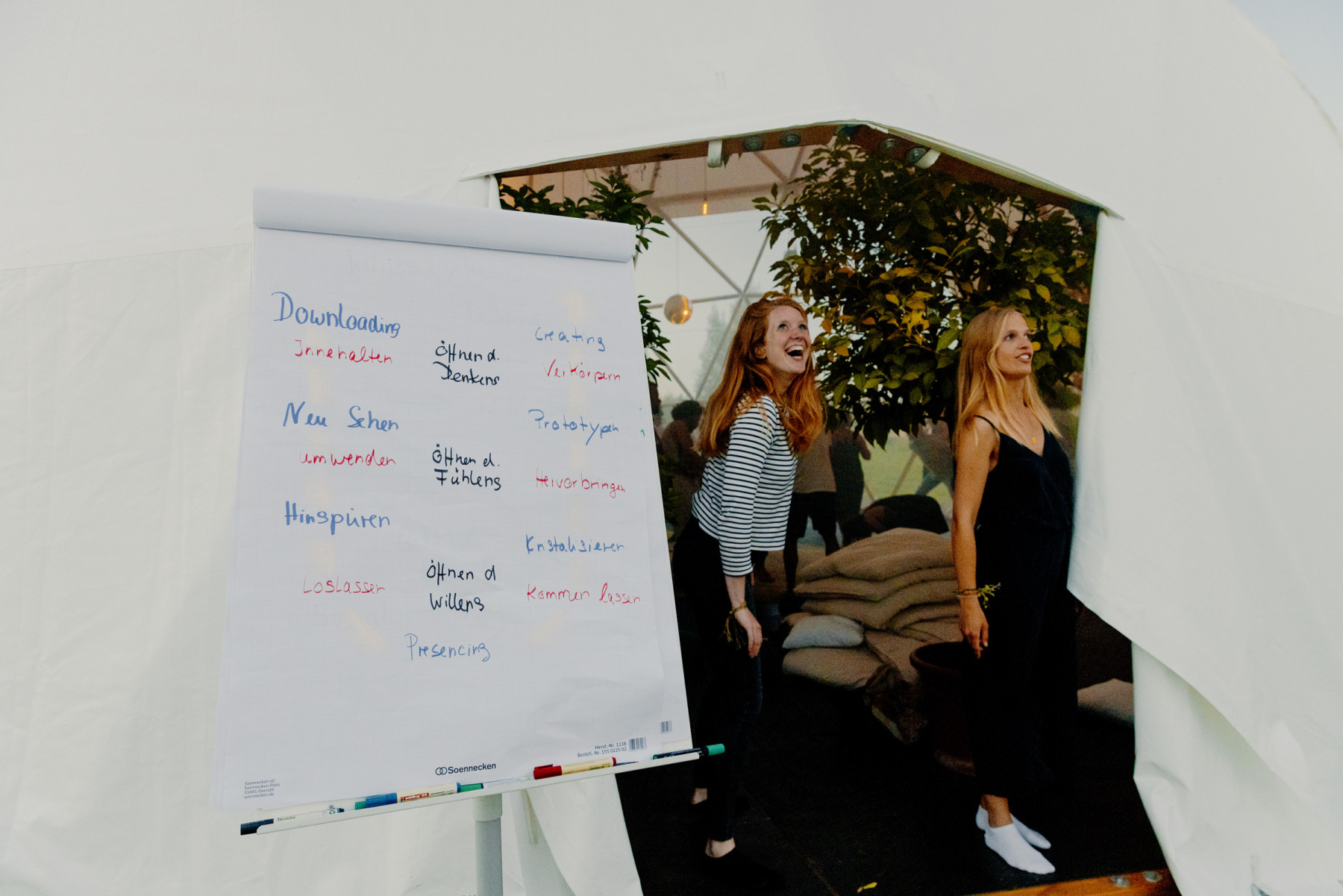 Zwei Frauen stehen in einem weißen Kuppelzelt neben einem Flipchart. Auf dem Flipchart sind Begriffe auf Deutsch und Englisch handschriftlich notiert. Im Hintergrund sind Pflanzen und Sitzkissen zu sehen.