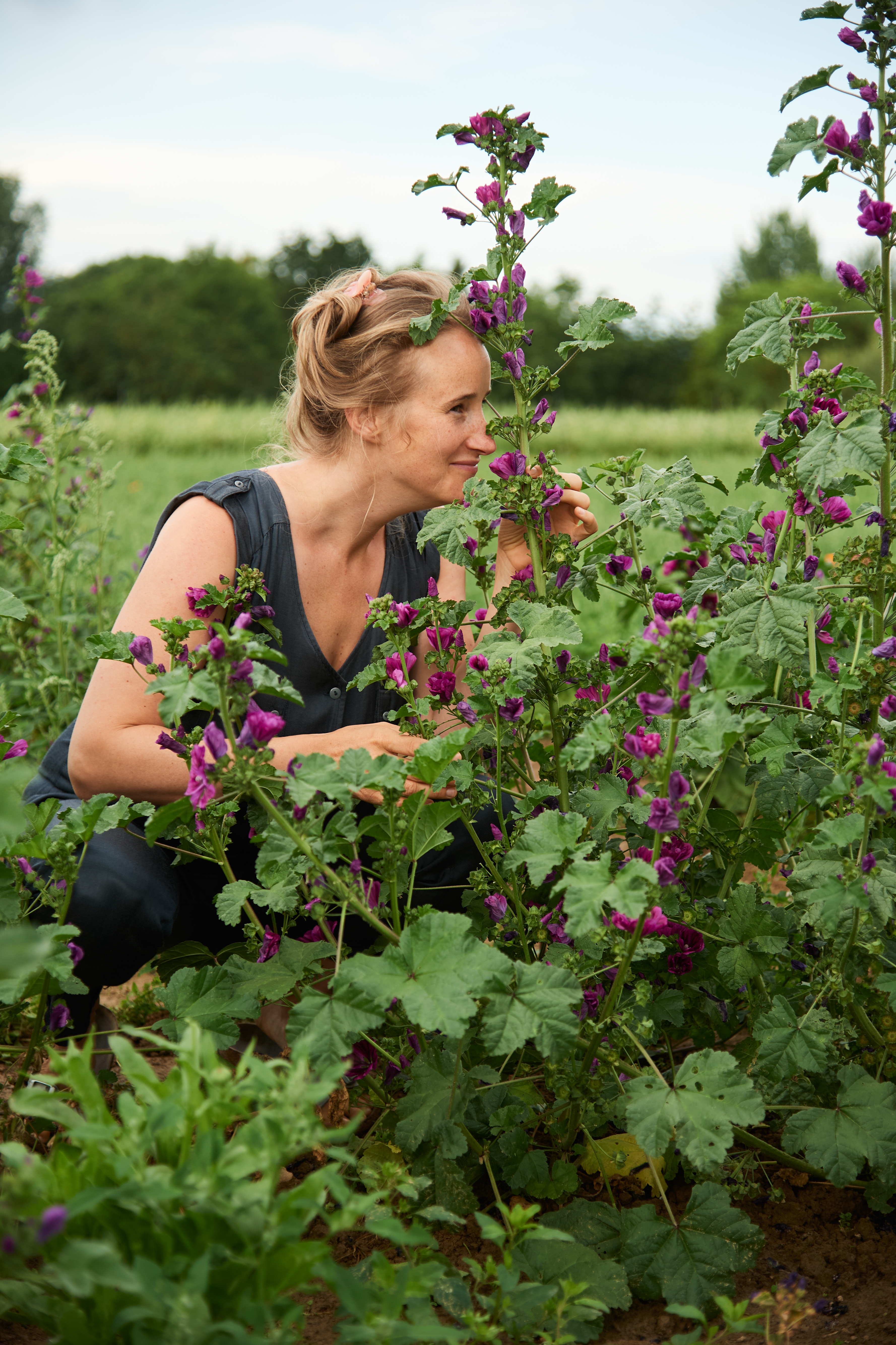 Eine Frau kniet auf einem Feld vor einer Malve und riecht an einer Bl&uuml;te. Sie l&auml;chelt.