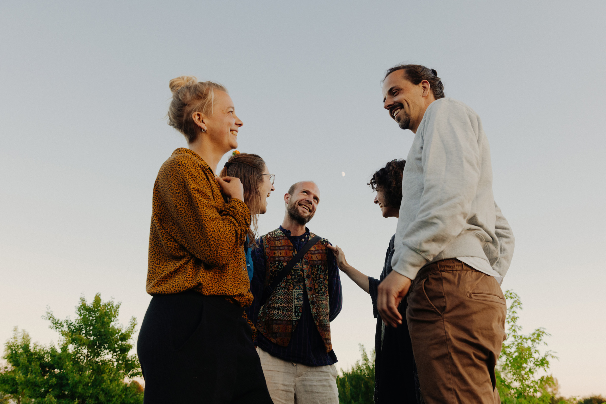 Sechs Personen stehen eng beieinander im Freien. Sie lachen und unterhalten sich. Die Stimmung ist leicht und offen, der Himmel dar&uuml;ber ist klar, mit zartem Licht.