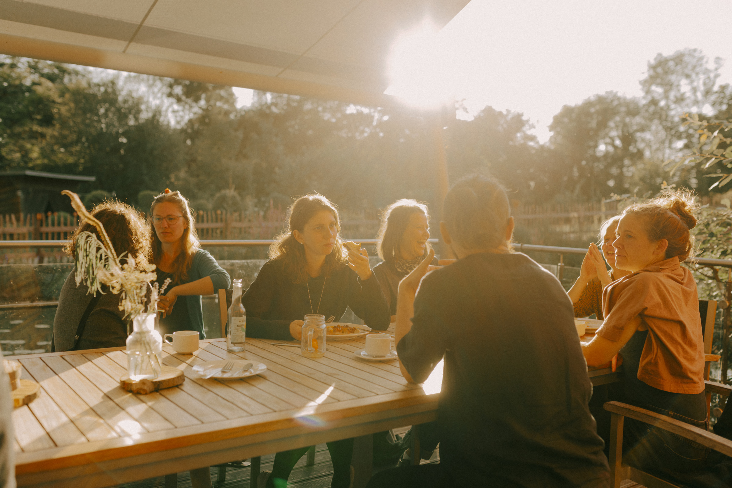 Eine Gruppe von etwa acht Personen sitzt an einem Holztisch im Freien. Die Sonne steht tief und wirft warmes Licht. Es wird gegessen, getrunken und sich unterhalten. Die Stimmung wirkt entspannt.