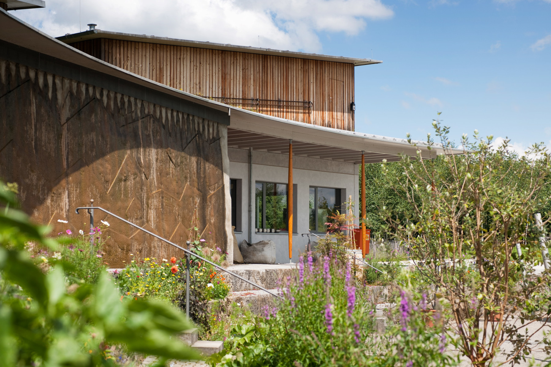 Modernes Gebäude mit Holz- und Lehmfassade, umgeben von blühenden Pflanzen und Gartenbeeten unter blauem Himmel.