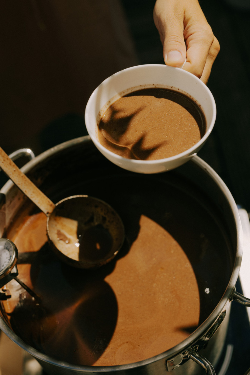 Ein großer Edelstahltopf ist mit heißer Schokolade oder Kakao gefüllt. Eine Hand hält eine weiße Tasse mit frisch eingeschenkter Portion.