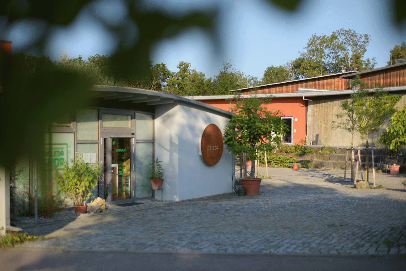 Ein kleiner, heller Pavillon mit Glastür und Pflanzen davor. Ein rundes Holzschild ziert die Wand. Durch Blätter im Vordergrund blickt man in einen ruhigen Hof mit Kopfsteinpflaster.