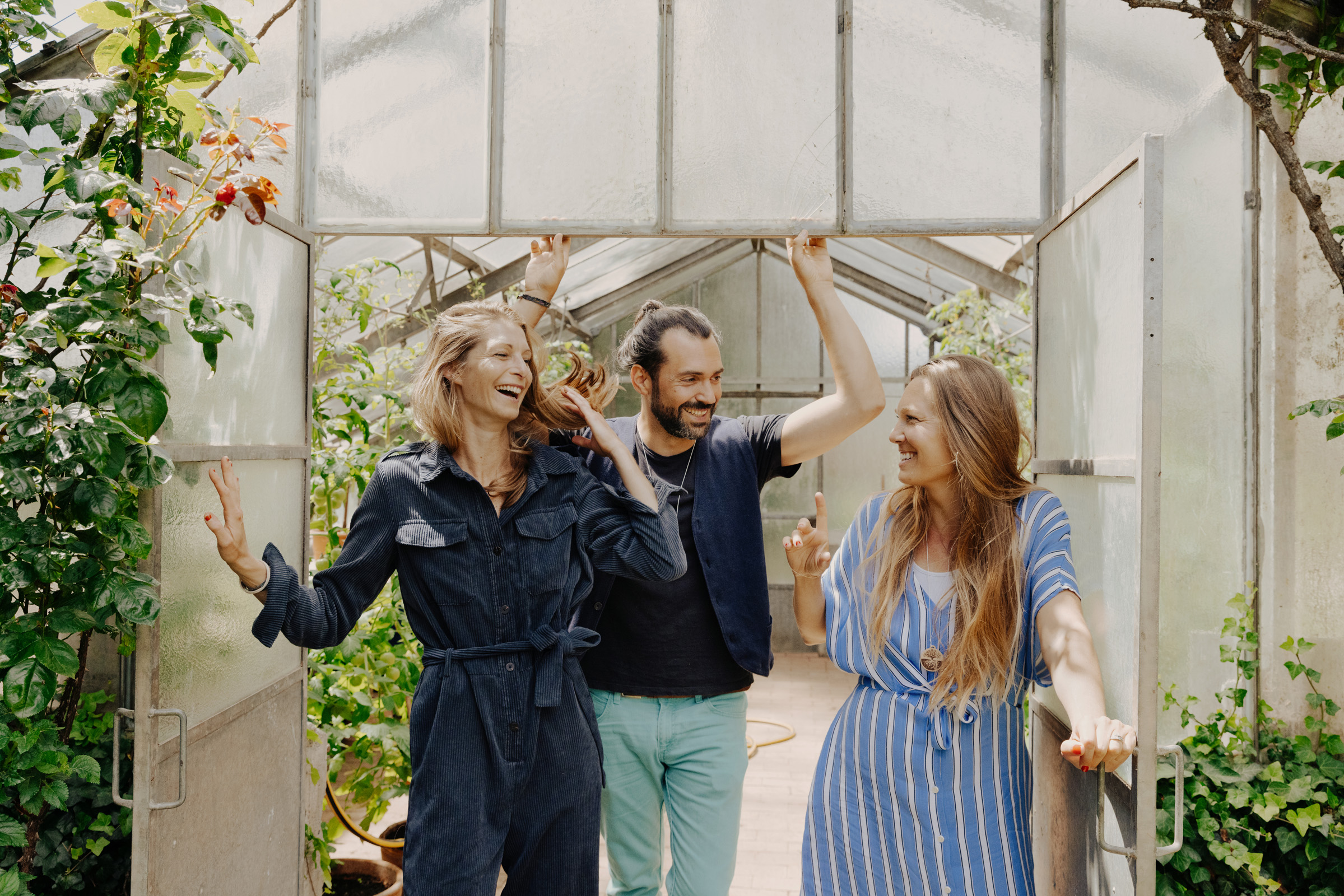 Drei lachende Erwachsene stehen am Eingang eines Gew&auml;chshauses aus Glas und unterhalten sich gut gelaunt zwischen gr&uuml;nen Pflanzen.