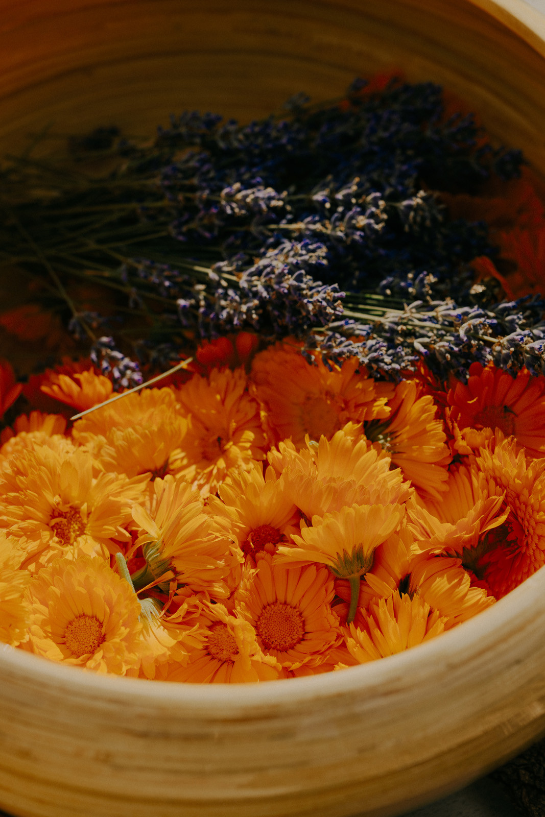 In einer runden, flachen Holzschale liegen leuchtend orangefarbene Ringelblumenblüten. Obenauf liegt ein gebündelter Strauß violetter Lavendelblüten. Das Licht ist warm und weich.