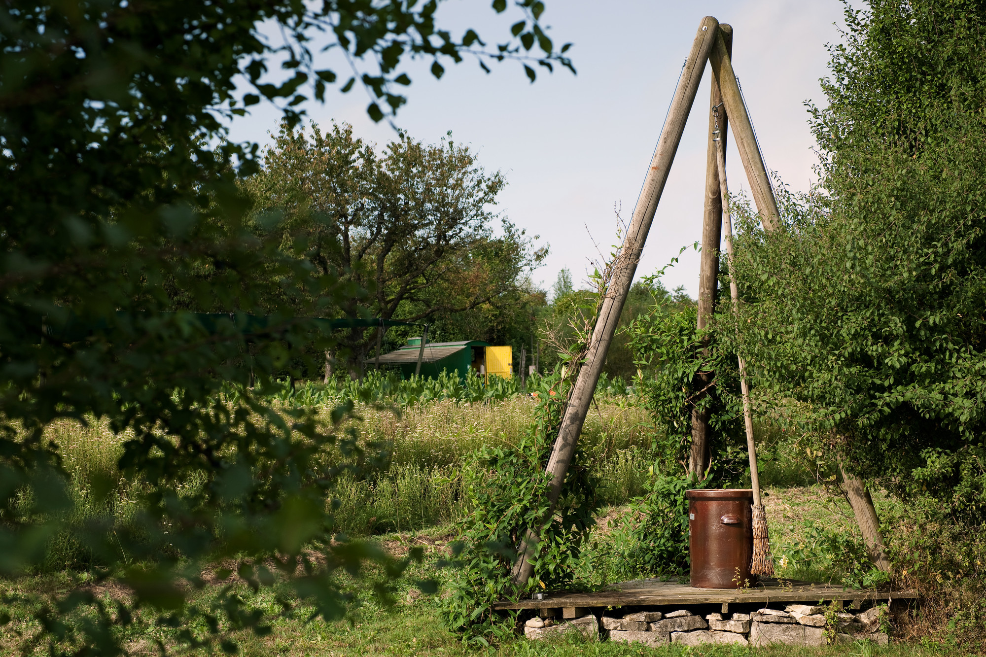Ein h&ouml;lzerner Dreibein mit Tonk&uuml;bel steht auf einer kleinen Plattform am Pr&auml;parateplatz inmitten einer gr&uuml;nen Wiese mit B&auml;umen und Str&auml;uchern.